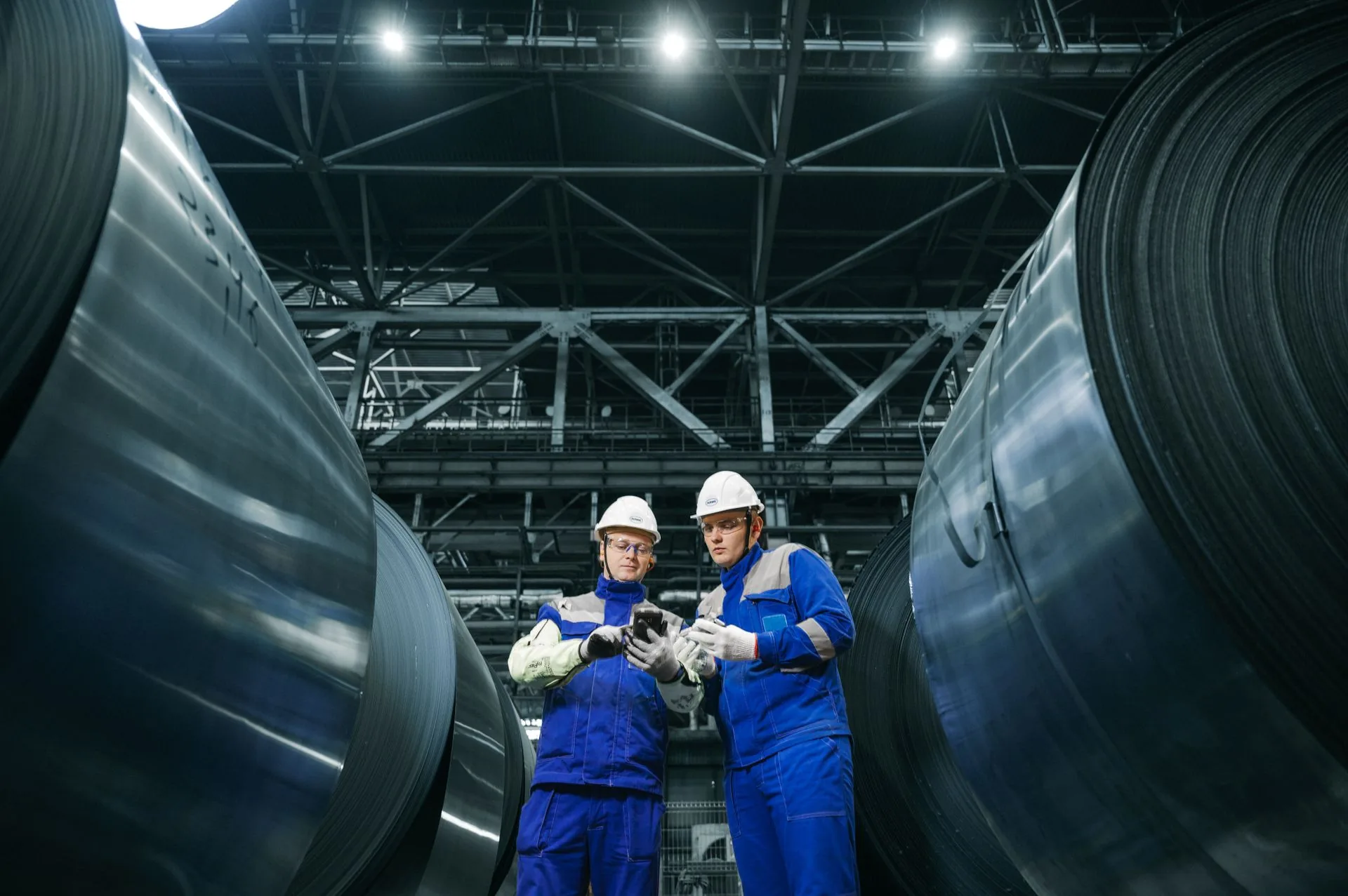 Dois profissionais com capacetes, óculos e luvas consultam um dispositivo em ambiente industrial.